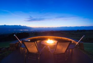 A lit fire pit with four chairs surrounding it, facing a great view of a valley at dusk. 