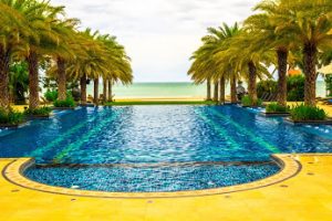 Outdoor swimming pool with palm trees lining it, facing the ocean. 