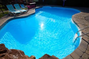 View of a clear blue swimming pool from above, surrounding by dark gray paving stones. 